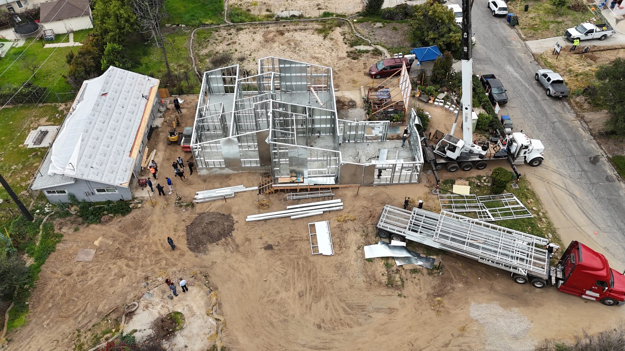 Aerial view of the 480 W Palm rebuild: factory-built light-gauge steel wall panels arriving by flatbed while the steel frame is assembled on the lot